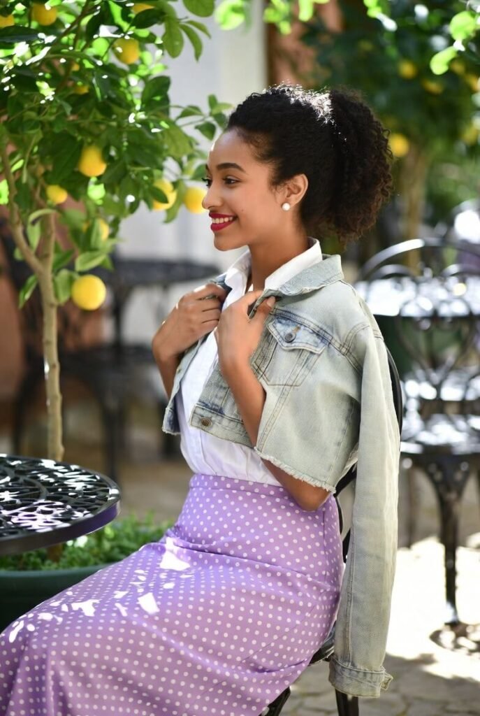 Young woman in a soft lavender polka-dot skirt, white blouse, and denim jacket seated at a garden brunch patio